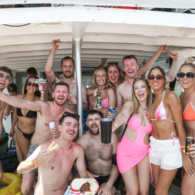 A group of people in swimwear are smiling and posing together under a boat canopy. They appear joyful, raising drinks and arms in celebration. The setting suggests a sunny day, likely on a boat or near water.