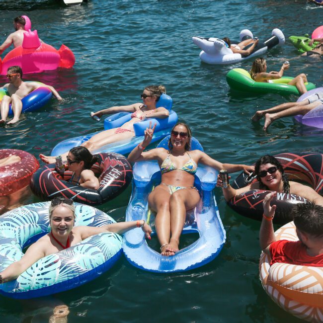 A group of people relax on various colorful floats in a sunny body of water. They are smiling and enjoying the warm weather, surrounded by vibrant inflatable rings and loungers. The scene is lively with a mix of beachwear and float designs.