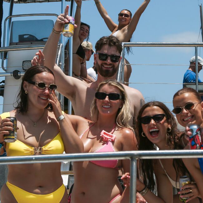 A group of people in swimsuits smiling and posing on a boat under a clear blue sky. The woman in front is wearing a pink swimsuit, surrounded by others in bright swimwear, holding drinks, with someone standing and waving in the background.