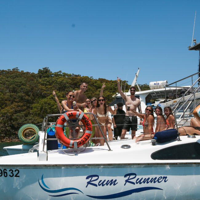 A group of people in swimwear are standing and sitting on a boat named "Rum Runner," waving and smiling. The boat is on calm water near a forested shoreline under a clear blue sky. Surfboards are visible on the boat.