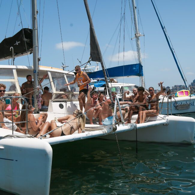 A group of people relaxes and socializes on two sailboats in a sunny harbor. Some are seated, while others stand or recline on the deck. The distant city skyline is visible under a clear blue sky.