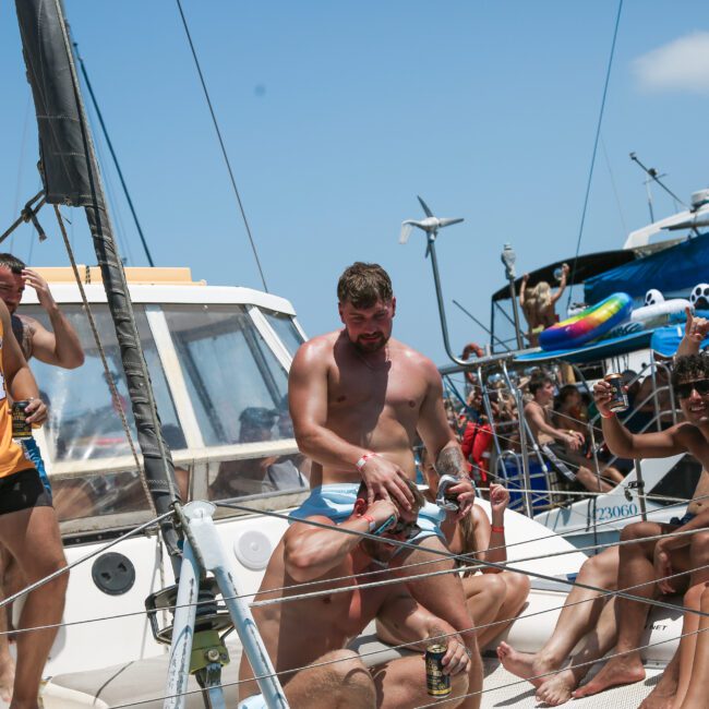 A lively group of people enjoying a sunny day on a sailboat. One person wears a Lakers jersey. The group is smiling, holding drinks, and surrounded by clear skies and other boats in the background.