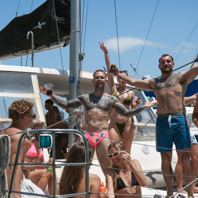 A group of people celebrating on a boat under a clear blue sky. Two men in the foreground are posing enthusiastically, one wearing a pink swimsuit. Other people are mingling and enjoying drinks in the background.