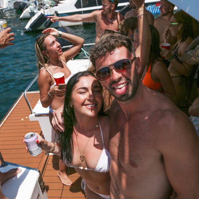 A group of people in swimsuits enjoying a sunny day on a boat. The scene is vibrant with socializing, and some are holding drinks. Other boats are visible in the background on a blue water body.