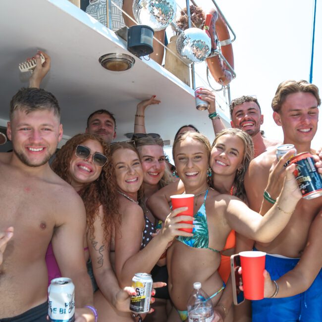 A group of young adults are enjoying a boat party, smiling and holding drinks. They are wearing swimsuits and seem to be celebrating under a sunny sky. The boat's deck and part of its structure are visible in the background.