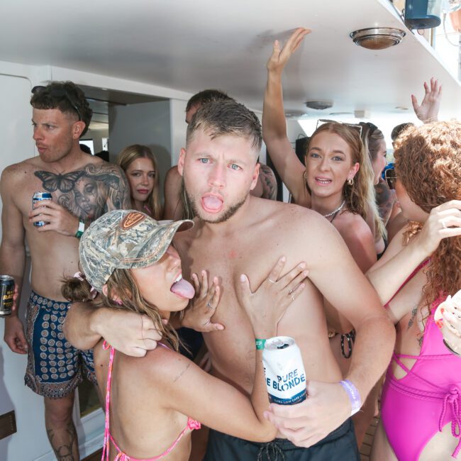 A group of people having fun on a boat party, with some in swimwear. A man in the center sticks out his tongue while a woman in a pink bikini playfully poses beside him. Other people are socializing and holding drinks.