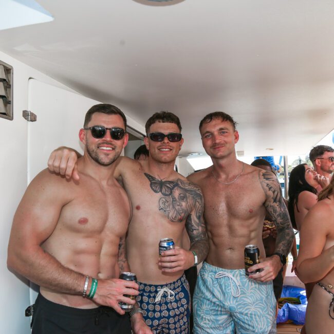 Three shirtless men wearing sunglasses pose together on a boat, smiling and holding drinks. They are surrounded by other people, some of whom are also holding beverages and enjoying the sunny day.