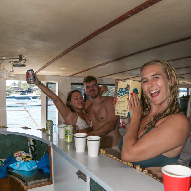 Three people smiling on a boat. The two in the background hold drinks, while the person in the foreground holds a box with the text "Tropic Growers." Disposable cups are on the counter in front of them. Outside, water and boats are visible.