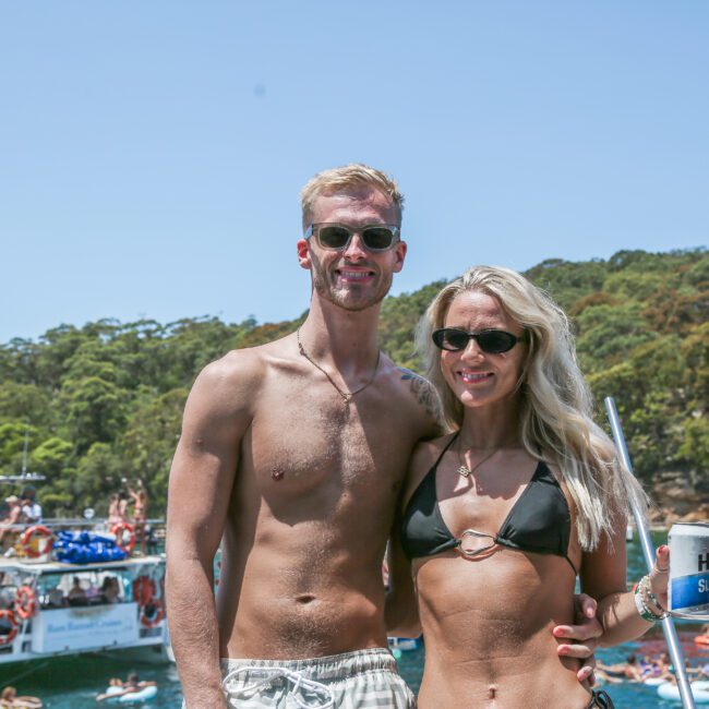 A couple wearing swimsuits and sunglasses smiles at the camera. They are on a boat surrounded by other boats and people enjoying the sunny day. Trees and a clear sky are visible in the background.