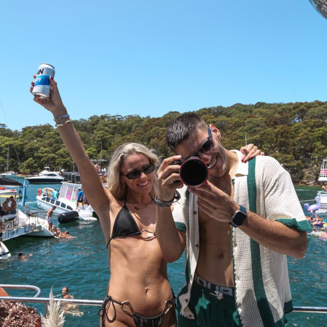 A man and woman pose on a boat. The woman in a black bikini raises a drink, and the man in sunglasses aims a camera. The background shows other boats and trees under a clear blue sky.