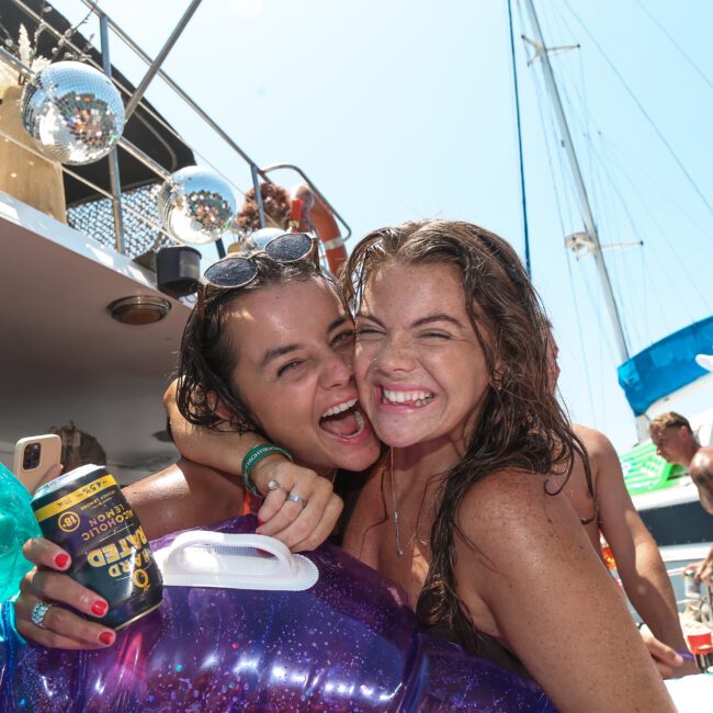 Two women smiling and posing together on a boat party. They are holding an inflatable float, surrounded by friends who are also enjoying the sunny day. A yacht and colorful decorations are visible in the background.