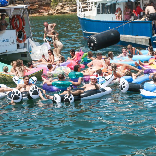 A group of people relax on inflatable pool floats in a lake, surrounded by boats. Some floats have animal paw designs. The scene is lively, with trees visible in the background and the sun shining brightly.