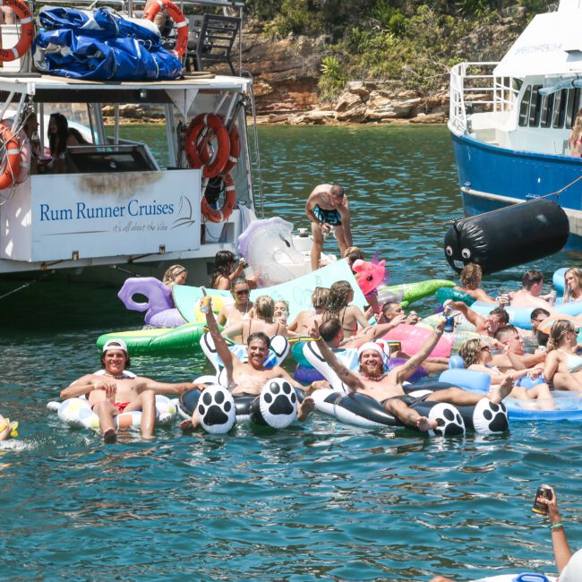 A lively gathering of people enjoying the water on inflatable floats, surrounded by boats. Most are wearing swimwear and appear to be celebrating in a tropical setting. The scene is festive with a mix of colorful floats and boats.