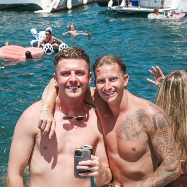 Two men pose happily on a boat, arm in arm with drinks in hand. They are both shirtless and smiling. Behind them, people are enjoying the water, with boats and an inflatable animal float visible.