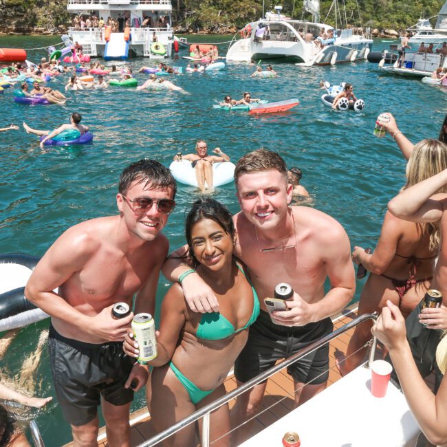 A lively boat party with people swimming and relaxing on inflatables in the water. In the foreground, three people in swimsuits smile at the camera, holding drinks. Various boats and partygoers are visible in the background.