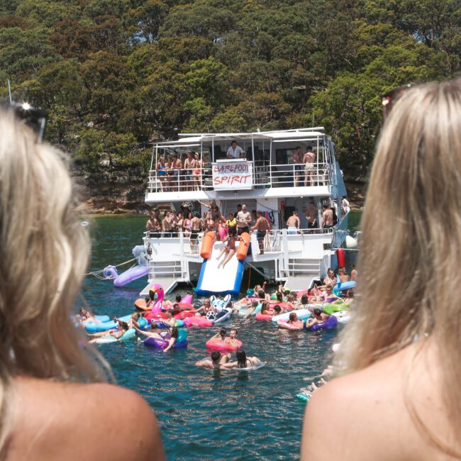 Two women observe a lively scene on a sunny day, where a crowd enjoys a boat party. People are swimming in the water and using inflatables near the multi-level boat. The backdrop includes lush green trees.