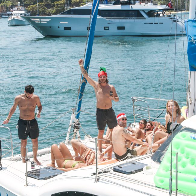 A group of people enjoying a sunny day on a sailboat, with one person standing and raising an arm in excitement. Others are lounging around and relaxing. A large yacht and green trees are visible in the background.