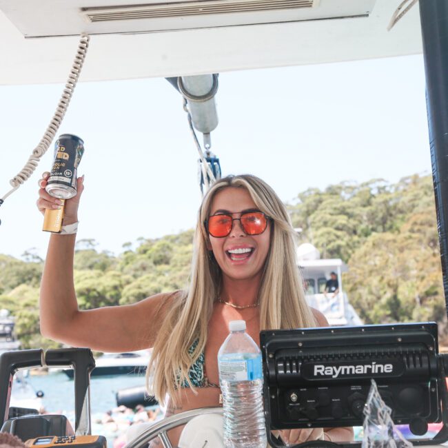 A smiling woman wearing sunglasses and a bikini top stands at the helm of a boat. She holds up a beer can in one hand. There is a water bottle in front of her. Trees and boats are visible in the background.