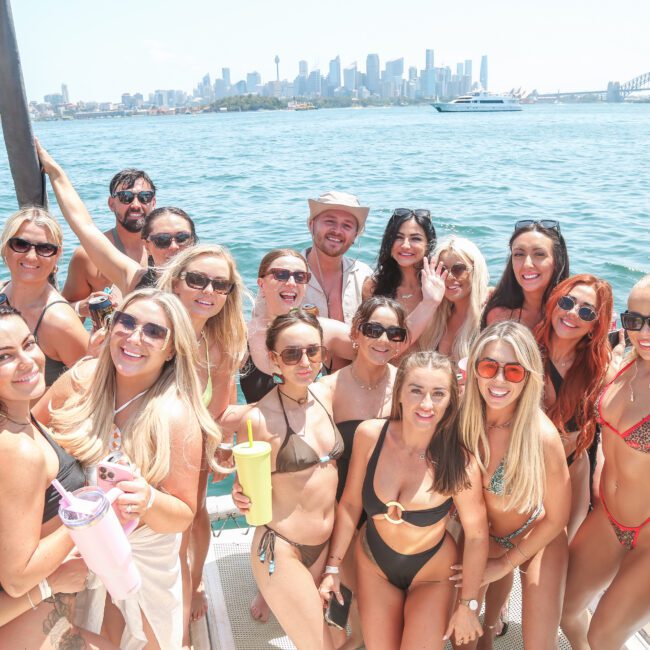 A group of people enjoying a sunny day on a boat, wearing swimsuits and sunglasses. They are smiling and posing for a photo with water and a city skyline in the background.