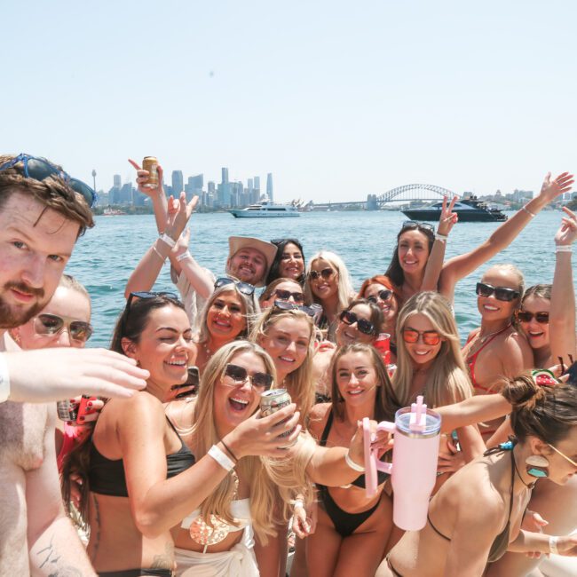 A group of people in swimwear smile and pose for a photo on a boat. They are enjoying a sunny day near the water, with a city skyline visible in the background. Some are holding drinks and wearing sunglasses.