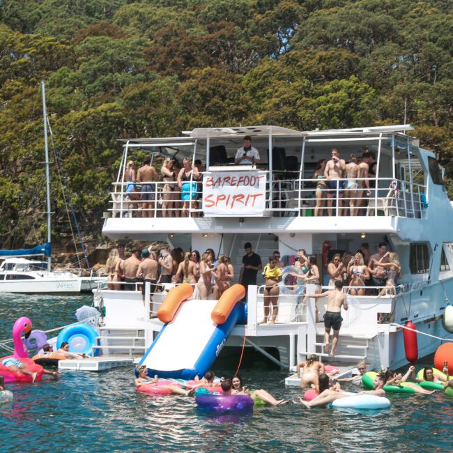 A crowded boat party with a "Barefoot Spirit" banner, moored by a lush, forested shoreline. People are enjoying the water, using colorful inflatables and inflatable slides, amidst a festive atmosphere. A distant sailboat is visible in the background.