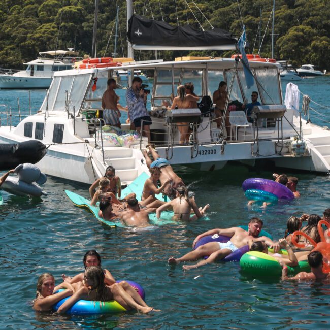 A group of people enjoying a sunny day on a catamaran and in the water. Several individuals are lounging on colorful inflatable rafts and tubes, surrounded by other boats in a scenic, leafy waterfront area.