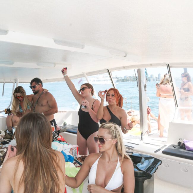 A group of people in swimsuits are enjoying a party on a boat. They are socializing and holding drinks, with some posing for pictures. Sunlight streams through the open windows, and bags and snacks are visible around them.