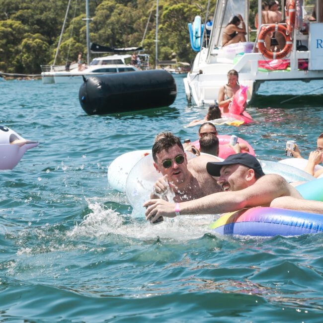 A group of people relax in colorful inflatable tubes in the water, near a boat labeled "Rum Runner Cruises." The scene is lively, with individuals enjoying the sunny day and water, surrounded by other boats and a green, tree-lined shore.