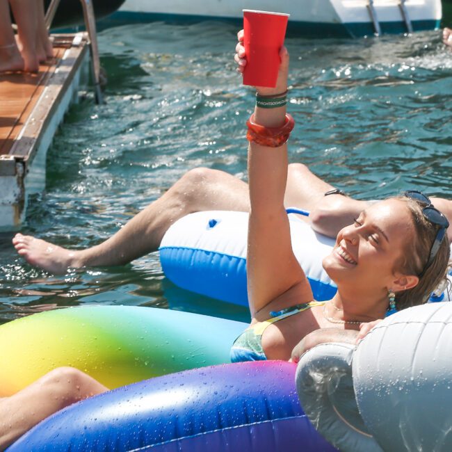 A person in a swimsuit is lounging on a colorful inflatable in the water, holding up a red cup and smiling. A boat is visible in the background, and another person's legs can be seen on a neighboring inflatable.