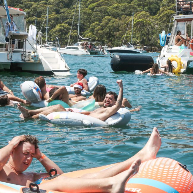 People enjoying a sunny day on a lake, floating on inflatable tubes and rafts. Several boats are anchored nearby with lush green trees in the background. Everyone appears relaxed and cheerful in the vibrant outdoor setting.