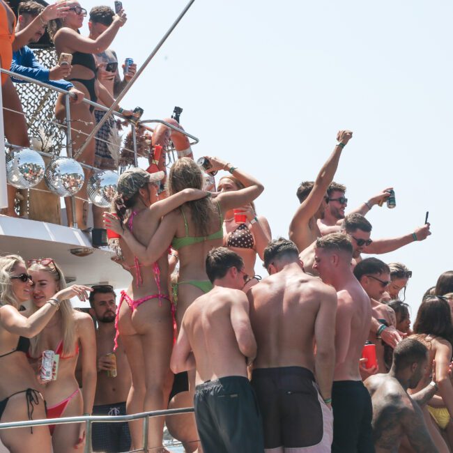 A lively group of people, mostly in swimwear, enjoy a party on a boat. Some are dancing, others are taking photos, and all appear to be celebrating under a sunny sky. The deck is crowded and festive.