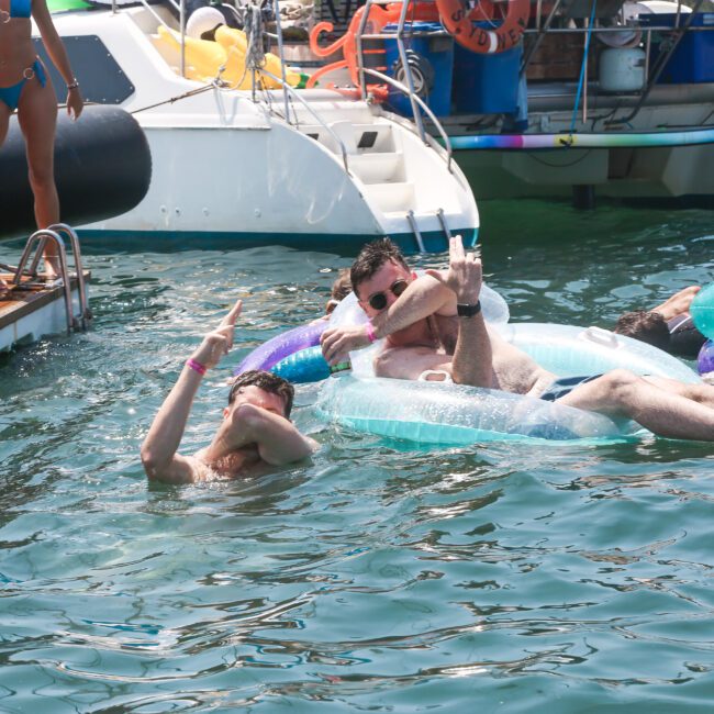 People enjoying a sunny day on the water near boats. One person is relaxing on an inflatable, while another swims nearby. The scene includes colorful floats and people on a dock, creating a festive and lively atmosphere.