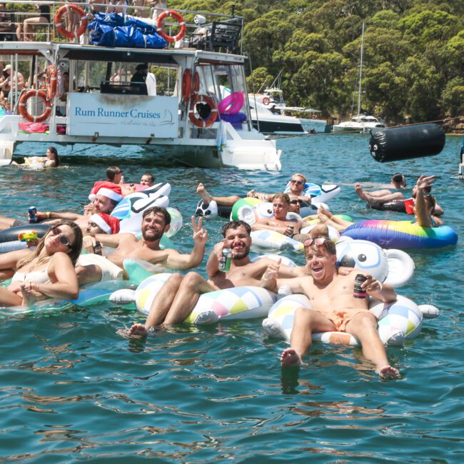 A group of people lounging on inflatable floats in a sunny water setting, surrounded by boats. Everyone is smiling and holding drinks, enjoying a festive atmosphere. Trees are visible in the background.