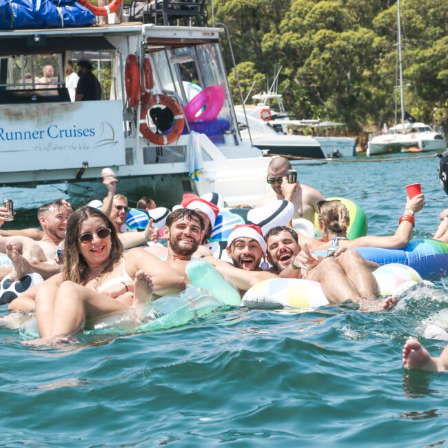 A group of people are joyfully floating on inflatable tubes in the water, with a boat and greenery in the background. They are dressed in swimwear and appear to be enjoying a sunny day at a gathering or party.