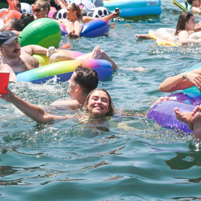 People enjoying a sunny day at a lake, swimming and floating on colorful inflatable tubes. One person smiles at the camera, surrounded by others relaxing on floats. A boat and more floats are visible in the background.