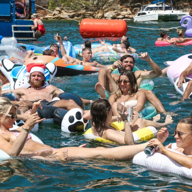 A large group of people enjoying a sunny day on a lake, floating on inflatable rafts and pool floats of various shapes. The atmosphere is festive, with some wearing hats, and others holding drinks. Boats are visible in the background.