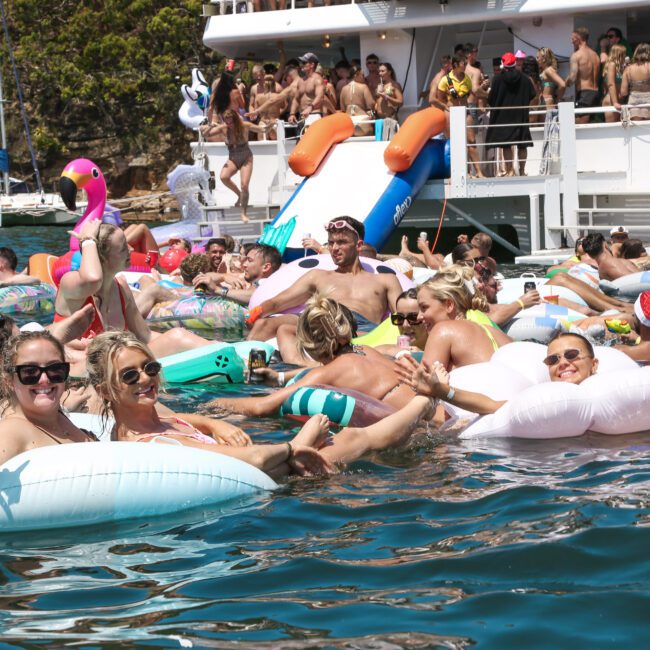 A lively group of people enjoying a sunny day on a lake, floating on colorful inflatables and inner tubes. A boat is anchored in the background, with more people gathered on its deck. It’s a festive, summery scene.