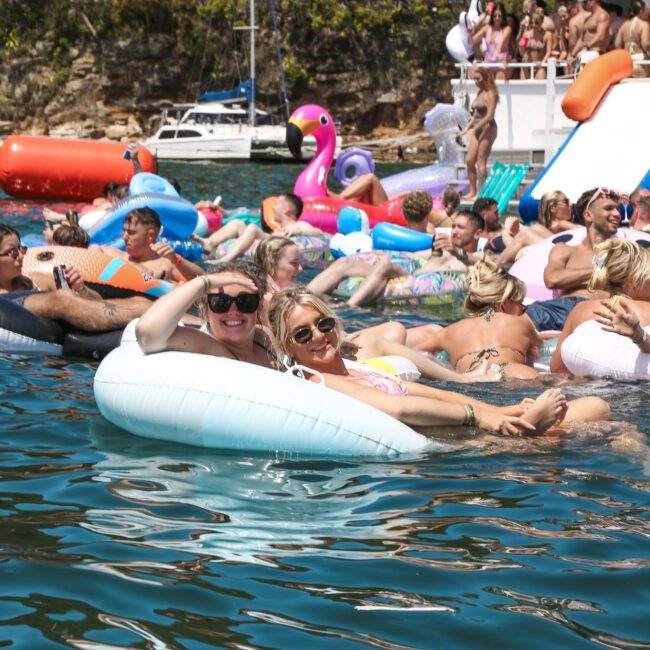 A lively scene of people relaxing on inflatable floats in a sunny lakeside setting. There's a slide from a boat where others are enjoying the water. Colorful floats include a flamingo and a Santa-themed float. Trees and a boat are visible in the background.