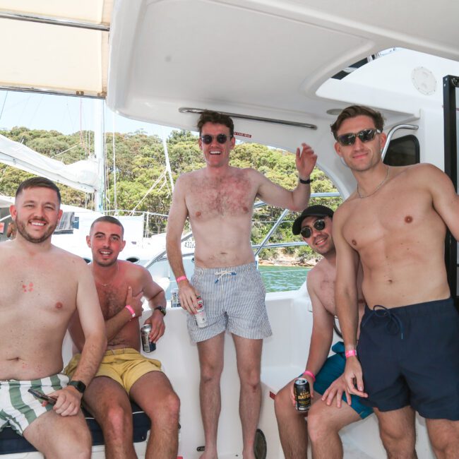 A group of five men in swimwear are smiling and posing on a boat. Some are holding drinks, and they're surrounded by a scenic view of trees and yachts in the background. It appears to be a sunny day.