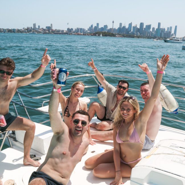 A group of six people is sitting on a boat, enjoying a sunny day. They are all wearing swimwear and smiling, some holding drinks, with the city skyline visible in the background. The ocean is calm, and another boat is seen in the distance.