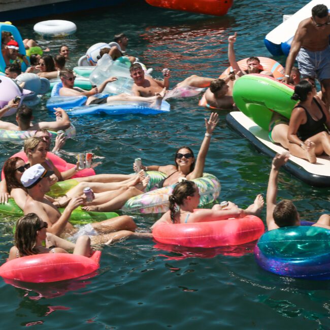 A lively group of people on colorful inflatable tubes and floats enjoy a sunny day on the water. Some are lounging, while others are engaged in conversation and waving. The scene is festive and relaxed, surrounded by clear, vibrant water.