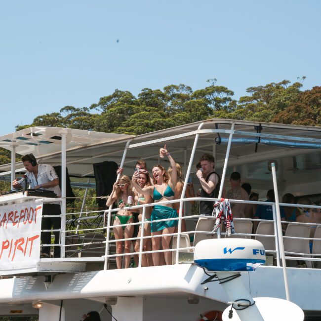 A group of people stand on the deck of a boat named "Barefoot Spirit," enjoying a sunny day. Some are posing for a photo, with trees and blue sky in the background.