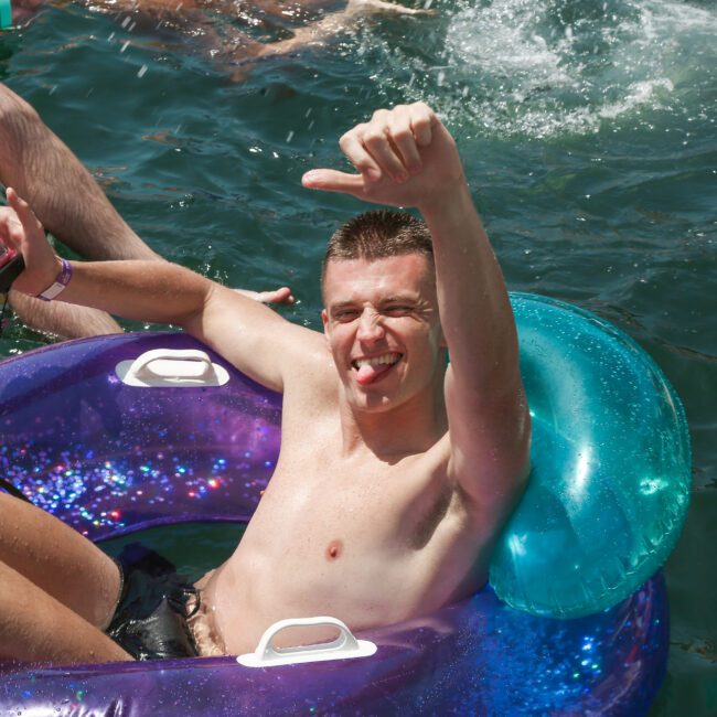 A person sitting in a colorful inflatable tube, floating on water. They are smiling, sticking their tongue out, and giving a thumbs-up. Another person is partially visible in the background on a similar tube.