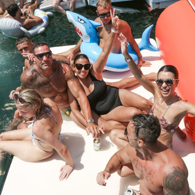 A group of people in swimwear enjoy a floating platform and inflatable shark in a body of water. Some are holding drinks, and others are posing for the photo, with smiles and peace signs.