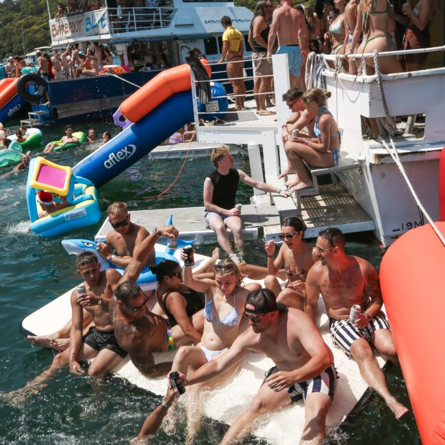 A group of people enjoy a lively boat party, some sitting on a floating platform in the water and others on the boat. A person is mid-dive into the water. Inflatable toys and a second boat are in the background.