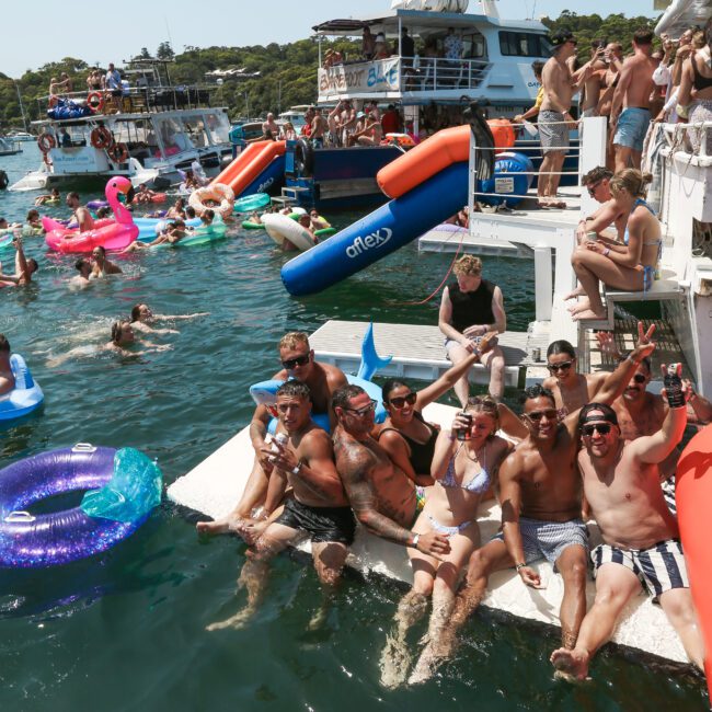 A group of people enjoying a lively party on boats and inflatables in the water. They are laughing, holding drinks, and surrounded by colorful floaties in a sunny, festive atmosphere.