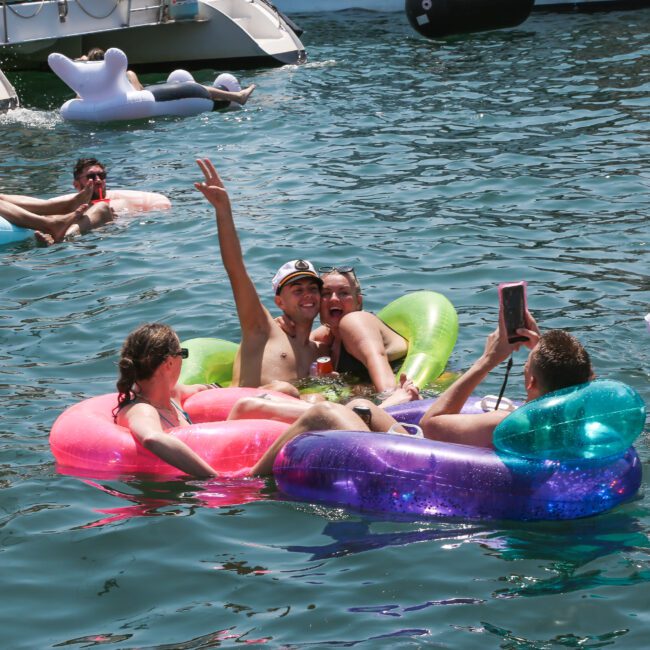 A group of people enjoy a sunny day on colorful inflatables in the water. Some are taking selfies, while others relax. Boats are anchored in the background. The atmosphere is cheerful and festive.