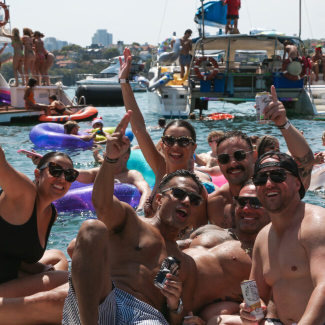 A group of people in swimwear enjoying a sunny day on the water, surrounded by inflatable floats. Boats and more people are visible in the background. Several individuals are posing with drinks and making peace signs.