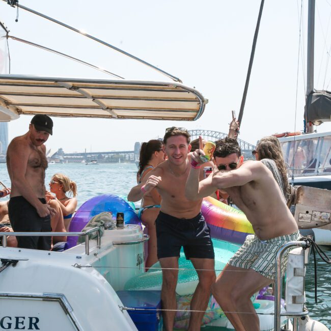 A group of people enjoying a sunny day on a boat. Two men in the foreground are playfully posing and smiling. The background shows other boats and a bridge in the distance. Various pool floats and drinks are visible.