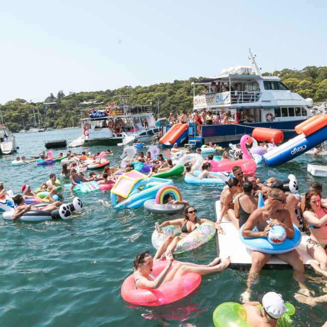 A lively scene on a sunny day with people enjoying a boat party. Many are on colorful inflatable floats, such as unicorns and flamingos, in the water. Boats are docked nearby, and the area is surrounded by lush greenery.